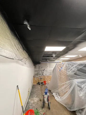 Vista del techo negro y sus paneles de iluminación sobre el interior protegido con plástico, con materiales y escalera al fondo.