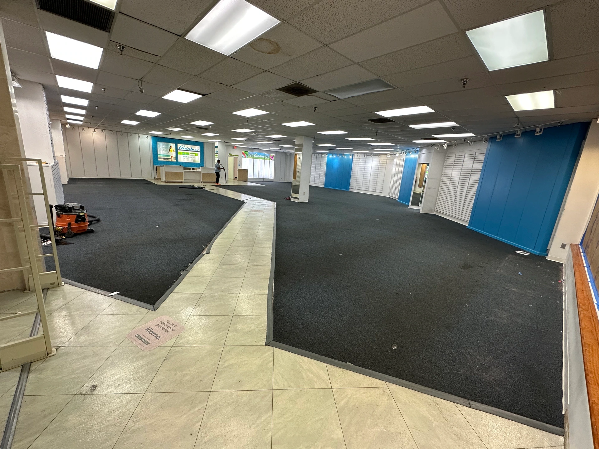Wide view of the existing space, with dark carpeted areas, light floor walkways, blue panels and an illuminated suspended ceiling.