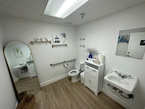 Bathroom with a white basin, mirror, toilet, grab bars and wood-look flooring.