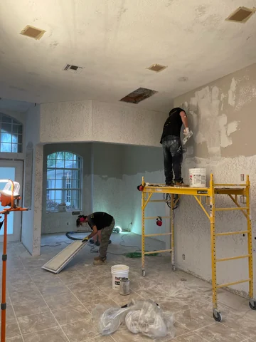 Two people working inside the home, one on a scaffold and another beside materials on the floor.