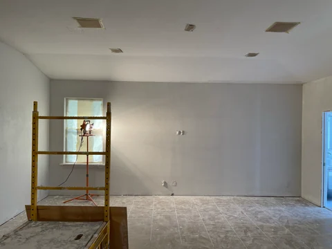 Wide view of a room with more uniform light-colored walls, a scaffold and a dust-covered floor during construction.