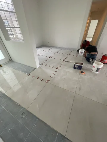 Workers installing light-colored floor tiles, with clips, wedges, buckets and tools visible.