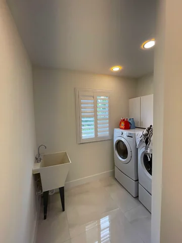 Laundry room with front-loading machines, a utility sink, white cabinets, a shuttered window and light-colored flooring.