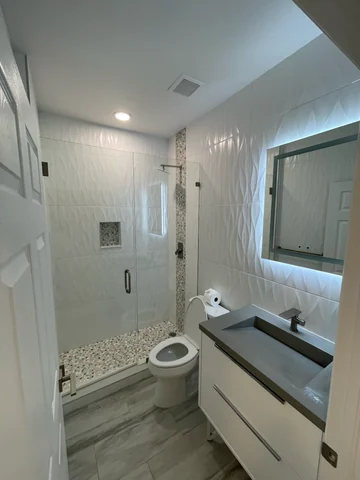 Bathroom with a shower behind a glass enclosure, light-colored wall finishes, a vanity and an illuminated mirror.