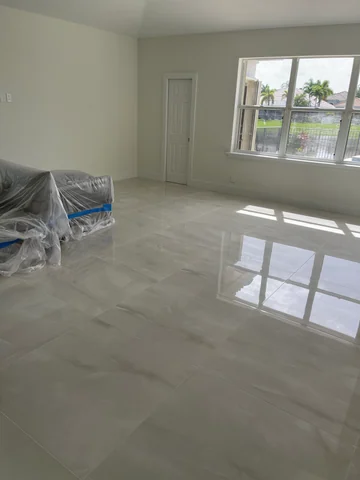 Living room with large light-colored floor tiles, a window facing the garden and a piece of furniture still covered for protection.