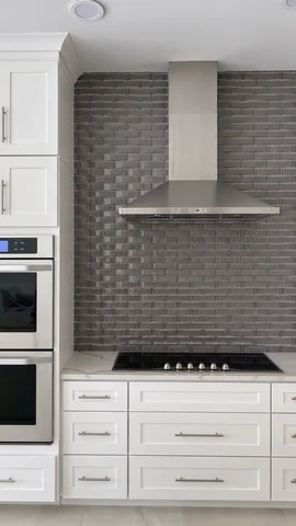 Kitchen detail with a metal range hood, gray backsplash, cooktop, ovens and white cabinets.