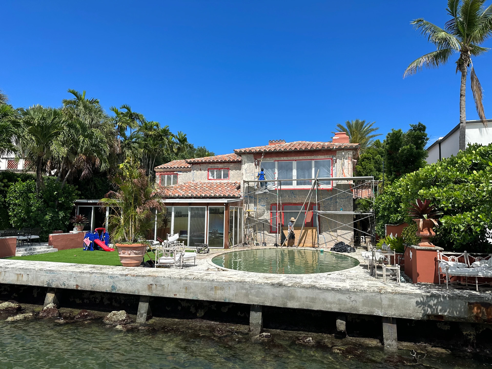 Wide view of a Miami Beach house beside the water, with a pool, palms and scaffolding along the rear facade.