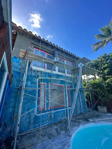 A worker on scaffolding in front of a blue and gray facade, with protected windows and a pool in the foreground.