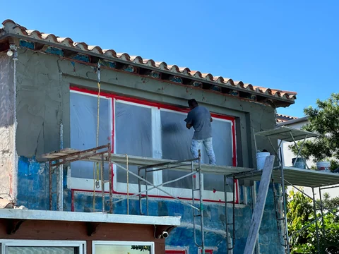A worker on a scaffold beside a protected window, with gray exterior coating and blue areas still visible.