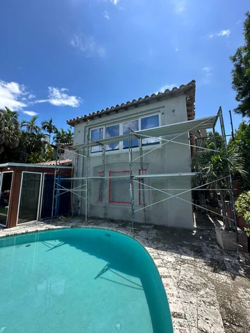 View from the pool toward a two-story gray-coated facade, with scaffolding and protected windows.