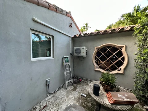 A courtyard between gray-coated walls, with a window, a circular diagonal-pattern opening and construction materials on the ground.