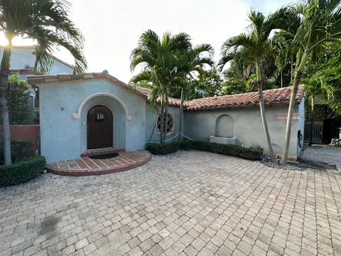 Wide view of the paved approach, arched front doorway, gray walls and palms around the house.