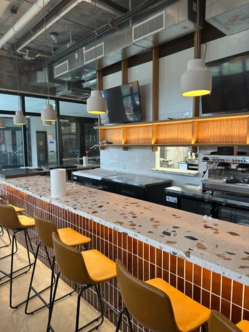 Closer view of the Craft bar with a speckled countertop, ceramic front, yellow stools and illuminated shelving.