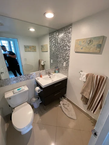 Existing bathroom with a white toilet, dark vanity, mirror and decorative wall finish behind the basin.