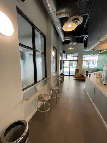 Circulación hacia la entrada de Smile Empanadas entre la barra junto a las ventanas y el mostrador, con banquetas e iluminación.